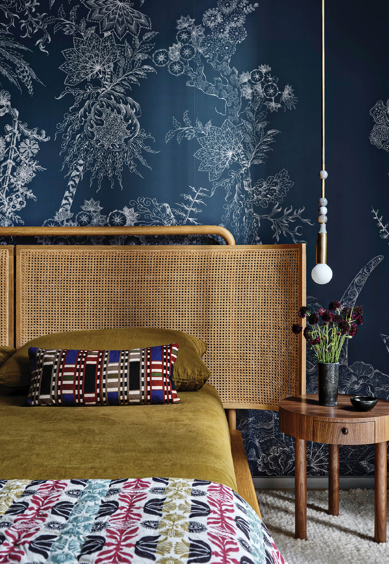 bedroom with dark blue patterned wallcovering, bed with cane headboard and walnut table