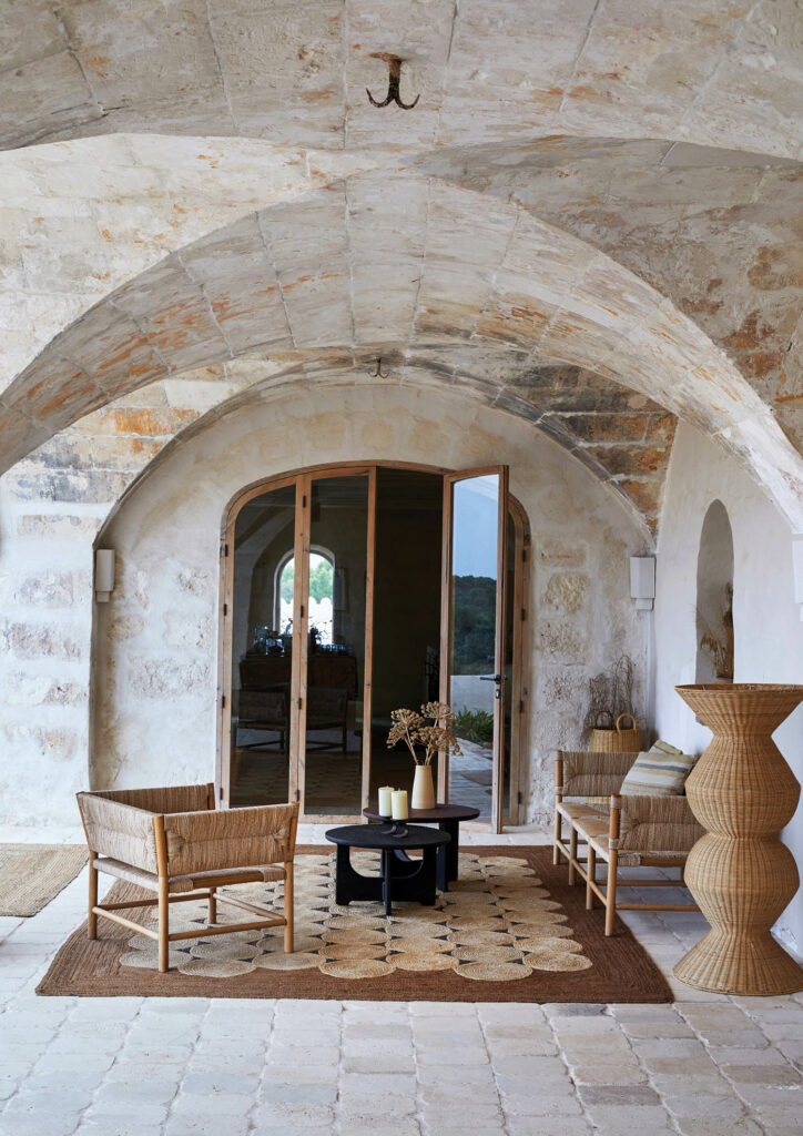 Restored limestone archways cover a terrace furnished with Radassié seating in oak and straw, wooden tables by Lucas Castex, and a Heaps & Woods Cronos lamp woven of sustainably sourced rattan.