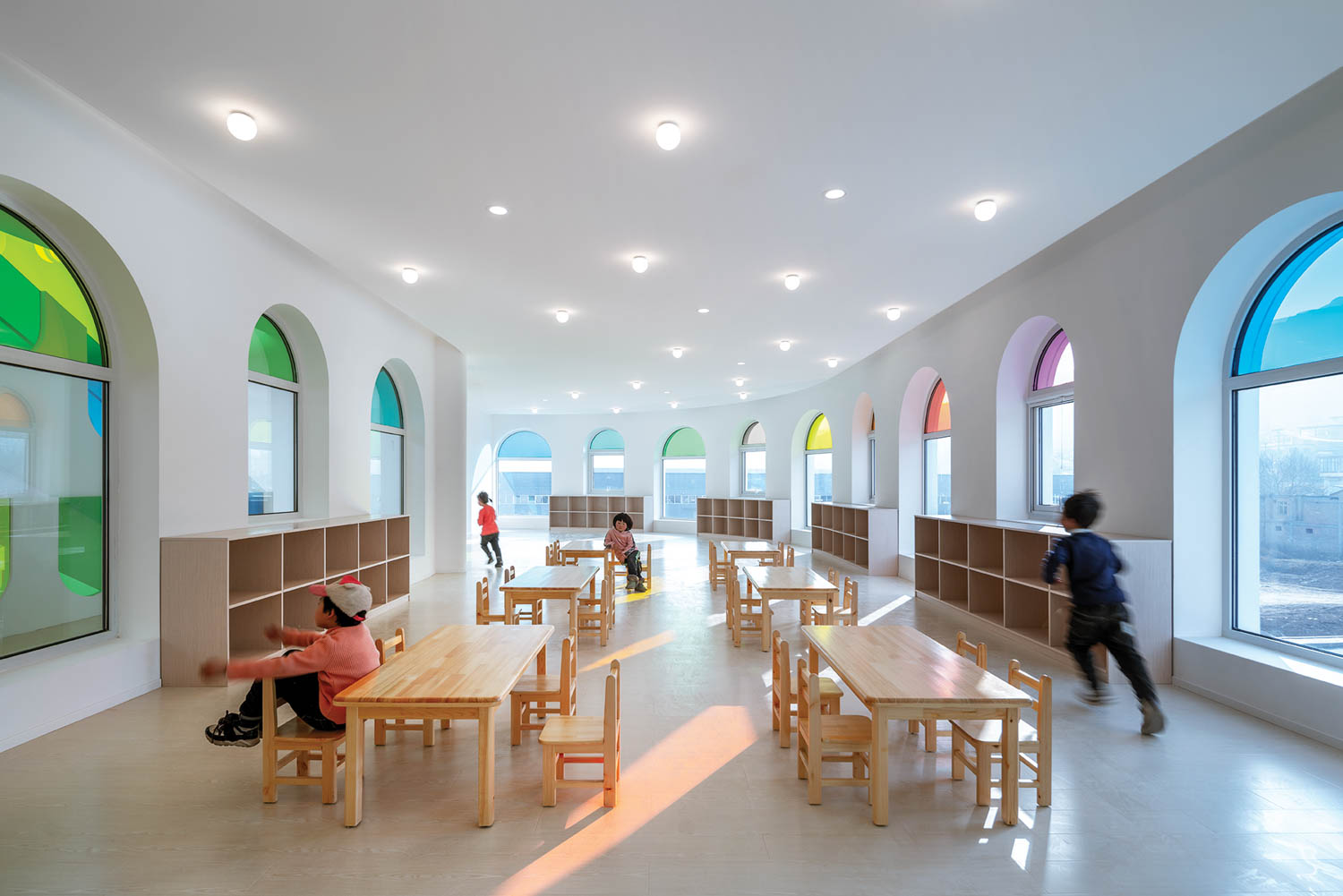 children sit at wood tables and chairs near arched windows