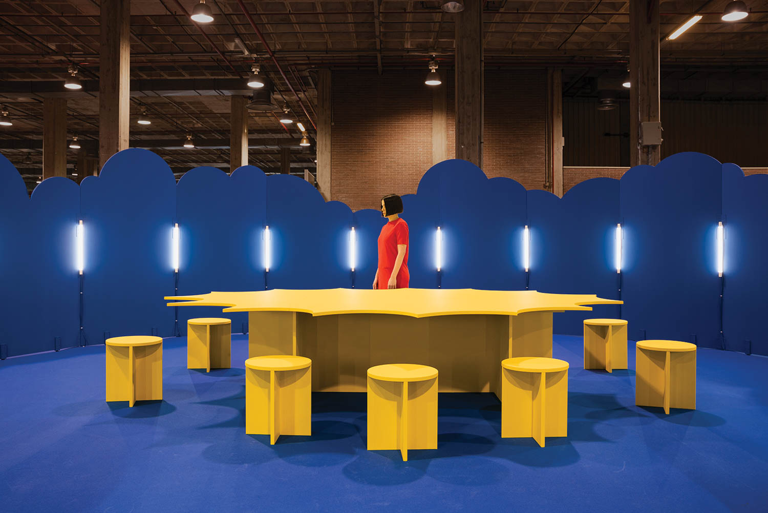 woman in bright red standing at a bright yellow table with chairs surrounded by blue clouds